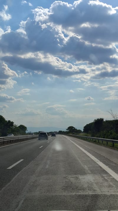 The image shows a view from a vehicle traveling on a highway in Tsaratsovo, Bulgaria. The highway has two lanes in each direction, separated by a median. The road surface appears to be asphalt, with white lane markings. There are guardrails on both sides of the road. The surrounding environment consists of trees and vegetation. In the distance, mountains can be seen. The sky is partly cloudy with dramatic cloud formations. Several cars are visible on the highway, all moving in the same direction as the viewer. The lighting suggests it is daytime, possibly late afternoon or early morning, given the quality of light and cloud shadows.