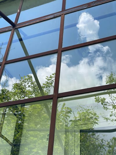 The image shows a close-up view of a windowpane with a grid of brown metal frames. The window reflects a bright blue sky with fluffy white clouds, as well as the green foliage of trees. The reflections are clear, suggesting a sunny day. The scene is photographed from a low angle, looking upwards through the window. The location is identified as Rudartsi, Bulgaria, though the image itself does not contain explicit geographical markers. It appears to be an exterior shot of a building, possibly a greenhouse or a structure with a large window surface. There are no people or animals visible in the image. The overall impression is one of nature and sky reflected on a man-made structure.