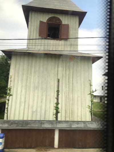 The image displays a tall, multi-tiered wooden structure, likely a bell tower or spire, situated in Vicovu de Sus, Romania. The upper section of the structure features a distinctive, slightly flared grey roof with dark brown trim, underneath which is an arched window with open brown louvered shutters. The main body of the structure is composed of vertical light-colored (pale off-white or light green) wooden paneling, exhibiting visible streaks and weathering. Approximately midway down the main section, a horizontal dark trim delineates a lower portion, below which the vertical paneling continues. Green vines or climbing plants ascend two vertical supports attached to the front face of the structure. The base of the structure includes a dark brown paneling section topped by a thick, dark grey horizontal band.

In the immediate foreground, multiple black utility wires run horizontally and diagonally across the frame, partially obscuring the view of the structure. The far right of the image is obstructed by a dark, checkered or mesh material, consistent with a window screen. A blue container is partially visible in the bottom left corner.

The sky above ranges from overcast light grey on the left to clearer blue with white clouds on the right. Lush green foliage is visible behind the left side of the main structure. In the middle-right background, a smaller, light-colored building with a sloped, metallic grey roof and a visible chimney stands amidst green grass, accessed by a dirt path. The overall lighting suggests daytime under mixed cloud conditions. No individuals are present in the scene.
