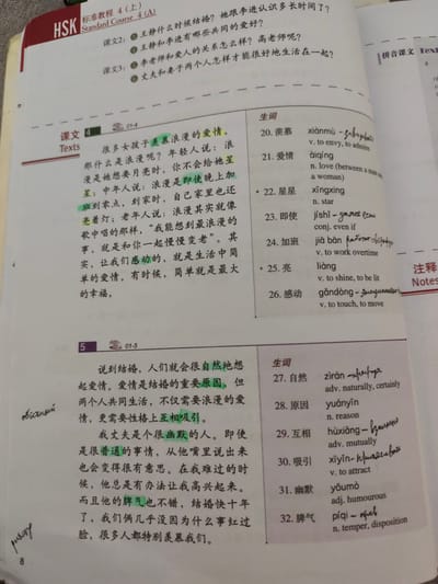 This is a close-up, eye-level shot of an open Chinese language textbook, likely from a classroom or study environment. The textbook is page 8 of a publication titled "HSK Standard Course 4 (上)". The page is predominantly white with black and some red text. There are handwritten annotations in blue ink, including pinyin transcriptions, English translations, and some Russian Cyrillic script, suggesting a learner is using the book.
The left side of the page contains lesson text for "课文 4" (Text 4) and "课文 5" (Text 5), with some passages highlighted in yellow and blue. The right side of the page lists vocabulary for the lessons, with Chinese characters, pinyin, and English definitions. Some of the pinyin and English definitions also have handwritten notes.
The environment appears to be indoors, with neutral lighting. There are no external environmental details visible that would indicate a specific location within Nanning, China. The focus is solely on the open textbook and its contents. The presence of other books or study materials is not visible.