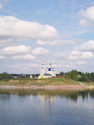 The image shows a yellow and blue church with golden domes standing on a grassy hill, reflected in the calm water below. The church is likely an Orthodox church, given the architectural style. The surrounding area includes trees and houses on the hilltop behind the church. The sky is blue with fluffy white clouds. The location is Глебово, Russia.