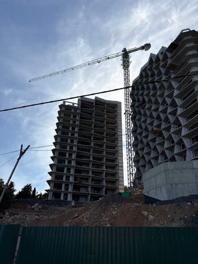 The image shows a construction site with two unfinished buildings and a crane in the city of Bondarenkovo, Ukraine. The building on the left is a tall, multi-story structure with a regular, rectangular facade. The building on the right has a more distinctive, angular design with a honeycomb-like pattern on its exterior. A large construction crane is positioned between the two buildings. The ground in the foreground is covered in dirt and rocks, with a green metal fence at the very bottom of the frame. Power lines are visible in the sky, and trees can be seen in the background on the left side of the image. The sky is partially cloudy.