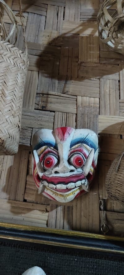 The image displays a multi-colored wooden mask against a woven bamboo wall in Cikanyere, Indonesia. The mask, positioned centrally, features a white base with prominent red, blue, black, and yellow painted accents. Distinct characteristics include large, round, red eyes with black pupils, a wide grimace revealing teeth, and two sharp, white fangs protruding from the upper jaw. A red marking is present on the forehead, complemented by blue lines above the eyes and yellow details on the lower face. To the upper left, a section of a light-brown, diagonally woven bamboo basket is visible, suspended. In the upper right, a partial view of a circularly woven basket is present. A dark, horizontally oriented frame with a gold trim traverses the lower foreground. Beneath this frame, a dark, textured surface is observed, with a small, pale, unidentifiable object partially visible at the bottom center. Shadows are cast upon the bamboo background, indicating an overhead light source. No individuals are depicted.