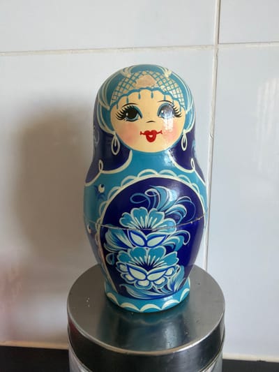 An unblinking, brightly painted matryoshka doll stands on a metallic cylindrical surface against a background of white tiled walls. The doll, predominantly blue and teal with accents of cream and red, is adorned with intricate floral patterns and a stylized human face. The doll's face features large, black eyelashes, blue eyes, rosy cheeks, and red lips. The tiled wall, with visible grout lines, suggests an indoor setting, possibly a kitchen or bathroom. The metallic surface on which the doll rests has a reflective quality, catching subtle light. The scene is static, with no observable action or interaction. The environment appears to be clean and well-lit. This image was taken in Sderot, Israel.