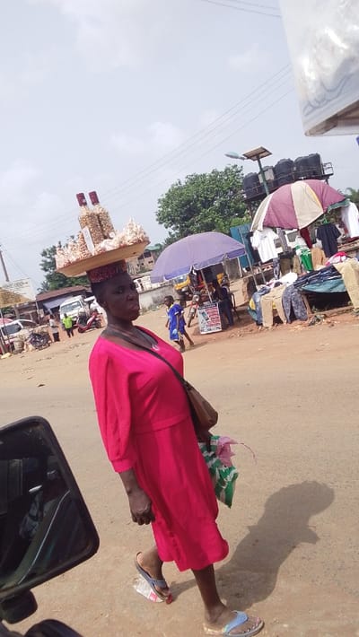 The image depicts a bustling street scene in Igbo Ukwu, Nigeria. In the foreground, an adult female with dark skin walks right-to-left across a reddish-brown, dusty road, her body angled slightly toward the viewer. She wears a vibrant pink, long-sleeved dress and blue-toned flip-flops; her left foot is seen stepping on a discarded clear plastic bottle. On her head, she expertly balances a light-colored wooden tray laden with goods for sale: several clear plastic bottles filled with light-colored nuts (likely groundnuts/peanuts) and numerous smaller clear plastic bags containing similar items. She carries a dark brown shoulder bag over her left shoulder and a striped green and white plastic bag in her left hand. Her facial expression is neutral. A distinct shadow extends from her onto the road surface to her right.

The background reveals a lively market environment. To the left, several buildings with corrugated roofs are visible, along with a white panel van, a red scooter, and multiple motorcycles. A street sign on a pole is partially visible. To the right, market stalls are set up under two large umbrellas, one solid purple and another striped purple and off-white, providing shade for displayed clothing items such as t-shirts and trousers. Several other individuals, including a child in a blue uniform, are present in this area. A yellow tricycle (keke Napep) is partially visible behind one umbrella. A white signboard with blue text stands near the stalls. Above the market setup, a tall structure supporting multiple black water tanks and a solar-powered light fixture is discernible. Green tree foliage is visible behind the stalls. The sky is overcast, and power lines traverse the upper portion of the frame. The lower-left corner shows a partial view of a dark vehicle mirror, suggesting the photograph was taken from inside or near a vehicle.