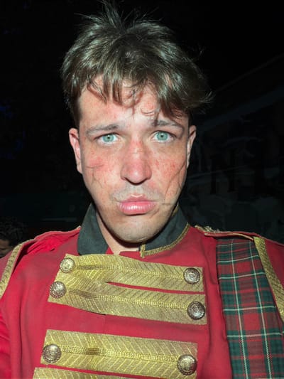 A man with blue eyes and light brown hair, his face smudged with red marks and what appear to be theatrical scars, is shown in a close-up portrait. He is wearing a red military-style jacket adorned with gold braiding and buttons, and a tartan scarf is visible on his right side. The background is dark and out of focus, with some indistinct shapes and lighting suggesting an indoor or night setting, possibly a performance or event. There are no discernible landmarks or elements that place this image specifically in Gollagutta, India.