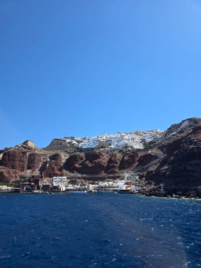 The image captures a stunning coastal view of a village built on a cliffside, overlooking a deep blue sea under a clear, bright sky. The village, characterized by its iconic white-washed buildings with blue accents, is nestled precariously on the steep, reddish-brown volcanic rock formations that characterize the landscape.
The scene suggests a sunny day, likely mid-morning or early afternoon, given the strong sunlight casting distinct shadows and highlighting the texture of the cliffs and the ripples on the water. The sea is a vibrant, deep blue, with small waves creating a dynamic, textured surface.
The architecture of the village is a prominent subject, with tightly packed houses cascading down the cliff. Elements like narrow pathways, balconies, and domed roofs are visible, hinting at the traditional Cycladic style of architecture. Some structures appear to be hotels or residences with swimming pools, suggesting a tourist destination.
There are no visible people or specific activities taking place. The focus is on the dramatic natural landscape and the human settlement integrated into it. The composition, with the vast expanse of sky on the left and the sea on the right, emphasizes the scale and grandeur of the location. The location cues, such as the unique cliff formations and the distinctive white architecture, strongly suggest Santorini, Greece, and specifically the village of Oia, known for its breathtaking views. The overall mood is one of serene beauty and awe-inspiring natural scenery. No text is visible in the image.