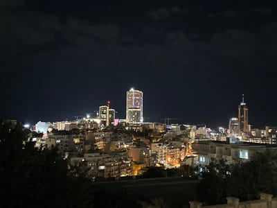 Nighttime cityscape featuring illuminated buildings. Skyscrapers punctuate the horizon, some with intricate light patterns. The foreground shows residential structures with lit windows, nestled among trees. A dark sky with hints of clouds provides a backdrop. Cranes are visible amidst the buildings, indicating ongoing construction. The scene is captured from a slightly elevated perspective.