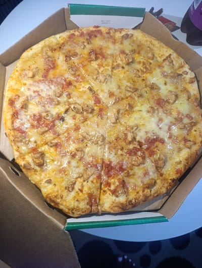 A pizza sits in a partially open cardboard box with a green border. The pizza is cut into slices and features a golden crust, a red tomato sauce base, melted cheese, and scattered pieces of what appears to be chicken. The box has an expiration date printed on the side. A bottle is partially visible in the top right corner, sitting beside the pizza box. The background surface is a light blue color.