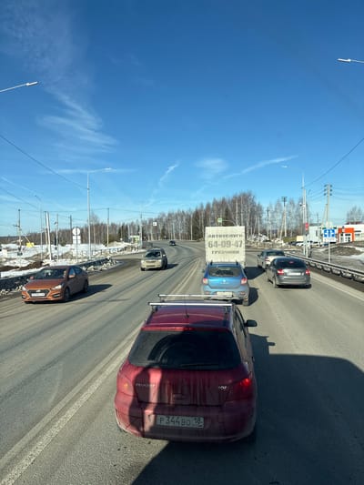 The image depicts a daytime traffic scene on a multi-lane asphalt road in Staroe Mikhailovskoe, Zav'yalovsky District, Udmurt Republic, Russia. The sky is clear blue with sparse white wispy clouds.
In the foreground, the rear of a dirty, dark red Peugeot 307 hatchback (license plate P344BO 18 RUS) is visible, equipped with roof racks. Directly ahead in the same lane is a light blue Dacia/Renault Sandero van (license plate P957TY 18 RUS) featuring a prominent white cargo box on its rear, labeled "АВТОУСЛУГИ 64-09-47 ГРУЗЧИКИ" (Auto Services 64-09-47 Movers). To the left of the van, in an adjacent lane, an orange Hyundai Solaris sedan (license plate P219ET 18 RUS) is driving alongside. Further down the right lane, a dark grey sedan (license plate X473BC 18 RUS) is observed, followed by a light grey SUV. All visible vehicle license plates bear the "18" region code, confirming the Udmurt Republic.
The road surface shows signs of wear and dirt, with clear white lane markings. Piles of melting snow and slush are accumulated along the road shoulders and median, indicative of early spring conditions. The surrounding environment consists of bare deciduous trees, possibly birch, lining the background. Numerous utility poles with overhead lines and modern streetlights with LED fixtures are present along the roadway. On the right side, an active red traffic light is visible, indicating a stop. Alongside it, blue and white road signs display a "СТОП" (STOP) symbol and an arrow indicating a mandatory right turn. A triangular pedestrian crossing sign is also visible. Further down on the right, a commercial building with an orange roof and white walls is situated. No specific individuals are clearly discernible in the vehicles or on the roadside.