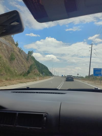 The image shows a view from the inside of a car looking forward on a road. The road is asphalt with white lines marking the lanes. There is a dark-colored car ahead, traveling in the same direction. To the left of the road, there is a steep, rocky hillside with some sparse vegetation. To the right of the road, there are power lines and a blue road sign with Greek text that appears to indicate distances to "Komotini" and "Nymfaia." The sky is blue with scattered white clouds. The dashboard of the car is visible in the foreground, partially obscuring the view. The scene suggests a journey on a highway or a main road in a semi-rural area.