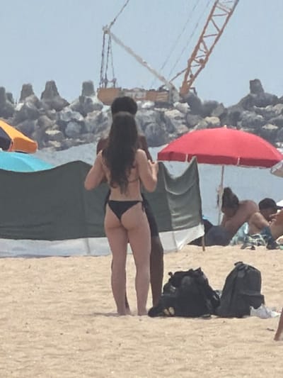 This is a photograph taken at the Praia da Baía in Espinho, Portugal. In the foreground, a woman in a black bikini is seen from the back, standing on the sand. To her left, a man's torso is visible. Behind them, a large green and white windbreak is set up. In the background, a red beach umbrella provides shade for people sitting on the sand. Further in the distance, a construction crane is visible against the sky, along with rock formations and the ocean. Two black backpacks are situated on the sand in the lower right portion of the image.