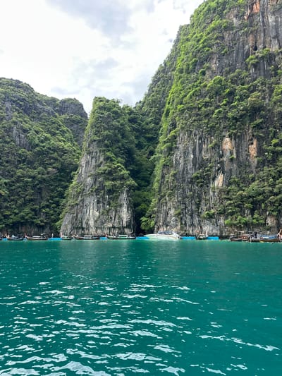 A photograph captures a turquoise body of water with ripples reflecting the overcast sky. Towering, rugged limestone cliffs covered in dense green vegetation rise dramatically from the water's edge, creating a dramatic natural landscape. A line of longtail boats, traditional Thai watercraft, are moored along the base of the cliffs. A few individuals are visible on or near the boats. The scene appears to be a coastal area, likely a bay or cove, with the geological formations suggesting a tropical environment. The image has the aesthetic of a tourist destination in Haad Yao, Thailand, known for its limestone karsts and clear waters.