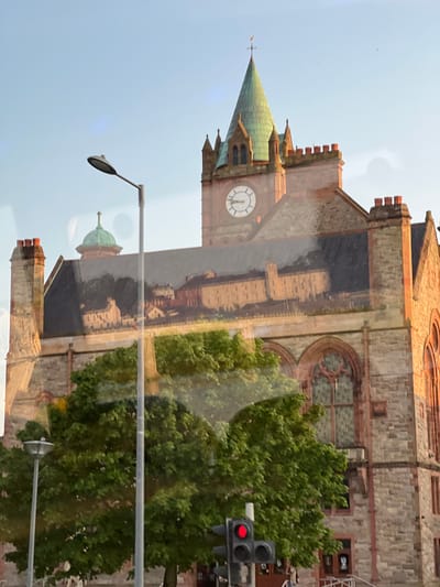 The image shows a reflection of a building with a clock tower and a green spire, possibly a town hall or a similar municipal building, superimposed on the actual building. The reflected image shows an older architectural style with multiple windows and a cityscape in the background. The actual building is made of stone and features red brick detailing around the windows. A large tree with lush green leaves partially obscures the lower portion of the building. In the foreground, a lamppost and a traffic light with a red signal are visible. The sky is clear with a pale blue hue, suggesting it might be morning or late afternoon. The overall impression is a layered image due to the reflection, creating a somewhat surreal effect. Without further information, it is difficult to pinpoint the exact location, but the architectural style suggests it could be a historic building in a European city. Based on the provided context of Derry/Londonderry, this building is likely the Guildhall in Derry/Londonderry, United Kingdom.