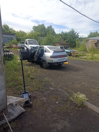 A silver sedan with its driver's side door open is parked on a dirt lot in a rural area, possibly in the city of Petukhovo, Russia. A blue hydraulic jack is positioned beneath the car's chassis. In the background, a white truck and a green sedan are also visible, along with some overgrown vegetation and a dilapidated brick building. The scene suggests an outdoor repair or storage area.