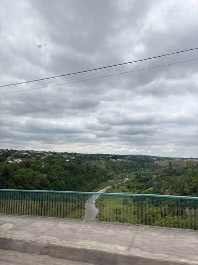 The image captures a panoramic view of a valley in Smotrych, Ukraine. In the foreground, a concrete barrier and a light blue railing, possibly part of a bridge or overlook, frame the scene. The railing exhibits a vertical stripe pattern due to reflections or the glass itself. Beyond the railing, a river meanders through a lush, green valley. Dense trees cover the slopes of the valley, with a small town nestled on the higher ground on the left side of the frame. The town features numerous houses with colorful roofs. The sky is overcast with gray clouds, suggesting a cloudy day. The overall impression is a serene landscape with signs of human habitation integrated into the natural environment.