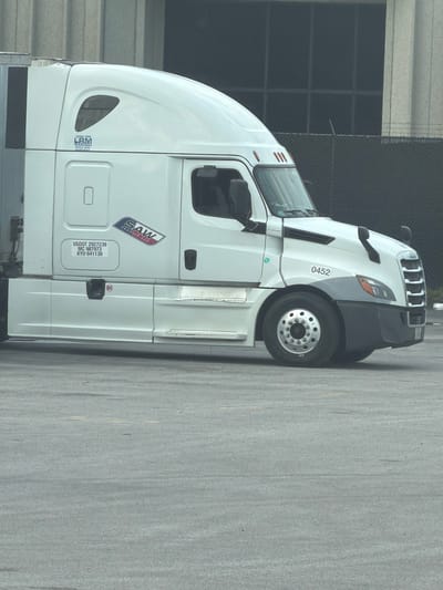 A white Freightliner semi-truck is parked in a lot. The truck has a "SAW TRANSPORT LLC" logo on its side, along with its USDOT number, MC number, and KYU number. The truck is also leased from "LBM LEASING." The scene appears to be in an industrial area, possibly a truck yard, and the background includes a building with large windows and a fence. There are no people visible in the image. The image does not contain any information to indicate its location in Chattanooga, United States of America.