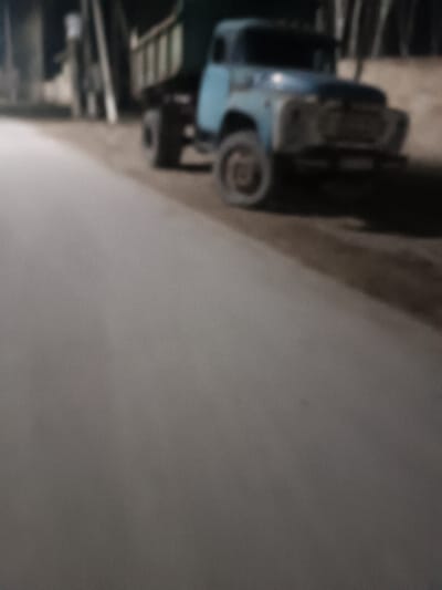 A blue truck is parked near a road. The truck, appearing old, has a green cargo bed. The foreground consists of an asphalt road, taking up most of the view. The background shows trees. The photograph's poor quality and low light cause details to be lost and make the image blurry.