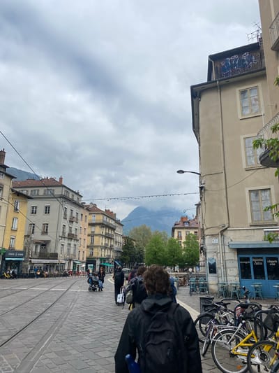 The image depicts a street scene in Grenoble, France, characterized by tall buildings, tram tracks, and a mountainous backdrop. The architecture features classic European facades with multiple windows and varying shades of yellow and gray. Tram tracks run through the paved street, indicating a central transportation route. Pedestrians are visible, including a person in a black jacket and a possible wheelchair user. Bicycles are parked, with one prominently yellow bike among others. Overhead cables and a street lamp cross the sky. Storefronts with signs, possibly for food establishments like "LOASIS KEBAB", add to the urban setting. The sky is overcast, suggesting a cloudy day. The mountains in the background provide a natural contrast to the urban environment.