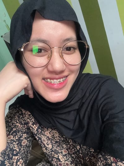 A close-up, chest-up image depicts a female individual in Patrang, Indonesia. She is wearing a black hijab and a dark garment with a multi-colored floral pattern. The subject is smiling, revealing upper teeth fitted with orthodontic braces featuring metal brackets and pink elastic bands. She wears round, gold-rimmed glasses, which exhibit a distinct green reflection on the left lens. Her left hand is positioned against her chin or cheek. The background consists of a wall with alternating vertical stripes of light green and white/cream. The scene is illuminated by an unseen light source.