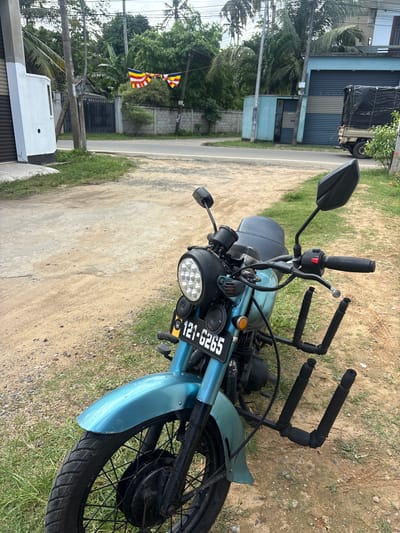 This image shows a close-up, eye-level shot of a light blue motorcycle parked on the side of a dirt and grass road. The motorcycle is the primary subject, with its front end and handlebars prominently featured. It has a black seat, a round headlight with multiple LED bulbs, and a license plate that reads "121-6265". A black, U-shaped rack is attached to the rear of the handlebars.
In the background, a paved road runs alongside the motorcycle. Further back, there is a blue building with what appears to be a shuttered storefront and a small blue booth. Trees, including palm trees, line the area, and there are electrical wires and poles visible. Two Buddhist flags, one with horizontal stripes of yellow, red, and blue, and another with diagonal stripes in similar colors, are hanging from a pole. The scene suggests a daytime setting under clear skies. The overall impression is of a quiet, possibly residential or commercial area in Kadewatta, Sri Lanka. No people are visible in the image.