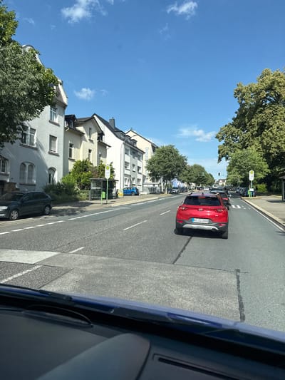 This image captures a street scene in Giessen, Germany, on a clear, sunny day. The perspective is from inside a vehicle, looking out at the road and surrounding buildings. The dominant subject is a red Kia SUV driving on the right side of the road, with a German license plate that reads "GI CI 2503". The car is in motion, as are other vehicles visible further down the street, indicating typical daytime traffic.

The setting is a residential and commercial street lined with trees and multi-story buildings. The architecture of the buildings suggests a European urban environment. One building, a light-colored apartment building, is prominently featured on the left side of the image, with visible windows and rooflines. There is also a bus stop with a shelter and signage visible on the right side of the road.

The time of day appears to be late morning or early afternoon, given the bright sunlight and the length of shadows. The sky is a vibrant blue with a few scattered white clouds, indicating fair weather. The overall mood is one of everyday life unfolding in a suburban or city neighborhood.

There is some visible text on signs and license plates. The license plate of the red Kia is legible. A sign with a "H" symbol, likely indicating a bus stop, is visible at the bus shelter. Text on a blue vehicle in the distance is difficult to read but appears to be advertising or signage.
