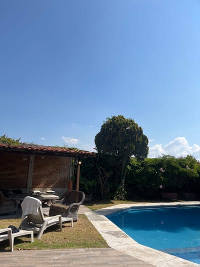 The image depicts a tranquil scene in El Oasis Valsequillo, Mexico, featuring a clear blue sky with scattered clouds above a residential property. The property includes a swimming pool with bright blue water, bordered by a concrete deck and adjacent green foliage. The scene incorporates outdoor furniture, including wicker and plastic chairs, partially situated on a grassy lawn that transitions into a tiled patio. A brick structure with a red tile roof sits to the left of the chairs. Lush greenery, including a large tree with pink blossoms, is present along the property line, adding a sense of privacy and seclusion to the space. No people are visible in the scene.