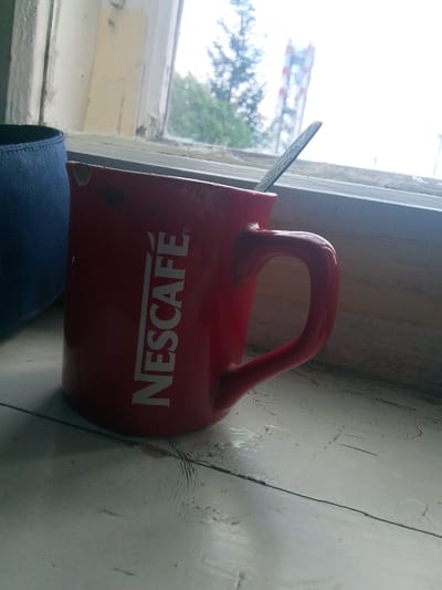 A red Nescafé mug with a spoon rests on a wooden windowsill. To the left of the mug, a dark blue fabric object is partially visible. Outside the window, trees and a red and white radio tower can be seen. The scene is likely in Volchansk, Russia, a town known for its industrial past and proximity to nature. The image captures a quiet moment, perhaps a break from work or a simple observation of the surroundings.