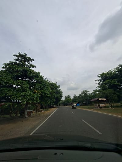 The image depicts a paved road lined with lush green trees on both sides. The sky is overcast with heavy clouds. On the left side of the road, there are several small wooden stalls or shops set up beneath the trees. On the right side, similar small structures are visible, some with awnings displaying text. A three-wheeled vehicle, commonly known as an auto-rickshaw or becak, is traveling down the road on the left side. The foreground shows the dashboard of a vehicle, indicating the photo was taken from inside a car. The scene suggests a rural or semi-urban setting in Glokaron, Indonesia, likely during daytime.