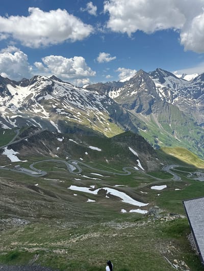 The image depicts a panoramic view of a winding mountain road in Fusch an der Großglocknerstraße, Austria, surrounded by snow-capped peaks and green alpine meadows. The road, a prominent feature, snakes its way up and down the mountainous terrain with multiple switchbacks. Several vehicles are visible on the road and in parking areas at different levels. Patches of snow are scattered across the landscape, even in the green areas. In the foreground, a small portion of a building with a grey, angled roof is visible on the right, and a person wearing a black and white outfit is standing in the lower left corner. The sky above is blue with scattered white clouds. The overall scene conveys the grandeur of the Austrian Alps and the engineering of the mountain road.