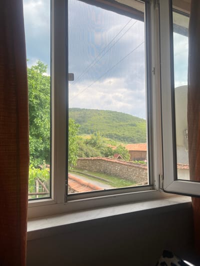 The image shows a view through an open window with a screen. The window overlooks a lush green hillside covered in trees, with a rustic stone wall and buildings visible on a path winding up the hill. The sky is cloudy and appears to be daytime. The foreground of the image shows the interior of a room, with wooden curtains framing the view. The overall impression is of a peaceful, rural scene, likely in a village or small town, given the architecture and natural landscape. The location context provided is Benkovski, Bulgaria. There is no visible text in the image.