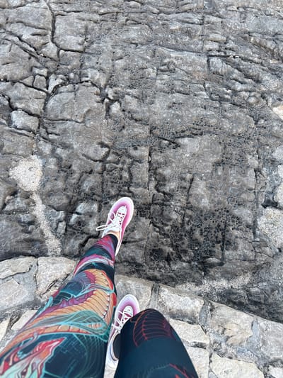 A top-down view captures the lower legs and feet of a person standing on a rugged, grey rocky surface in Seoca, Montenegro. The individual wears vibrant, multi-colored athletic leggings featuring an intricate, abstract organic pattern with hues of blue, green, orange, red, and purple on a dark background. One visible athletic shoe is predominantly pink and white with white laces, while the other is partially cropped. The rocky ground is highly textured, characterized by numerous cracks, fissures, and patches of small, circular indentations, possibly due to erosion or natural formations. Lighter grey rock sections are interspersed with darker, more weathered areas, and fine, light-colored granular material, likely sand or small pebbles, fills some crevices. The scene is brightly and evenly lit, suggesting daylight. No specific activity beyond standing or being present on the rocks is discernible, and there is no visible text.