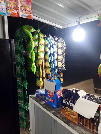 This media file depicts a roadside snack stall in Gampongpaya, Indonesia, likely at night given the artificial lighting. The stall is laden with an assortment of packaged snacks, hanging from the roof of a simple structure. Prominently displayed are bags of chips and crackers, with various brands and flavors visible.
Among the hanging snacks, several green bags labeled "Mini Stick" are clustered together. Other hanging items include bags with "Daia Floral" branding, and what appear to be potato chip bags labeled "POTATO". On the counter of the stall, several boxes of snacks are arranged. One box contains "Arden Choco Spekuloos" cookies. Next to it is a blue box of "OREO" cookies. There are also other packaged goods, including what look like biscuit rolls.
A bright, bare light bulb hangs overhead, illuminating the scene and casting a glow on the snacks. The background is dark, suggesting it is nighttime or the stall is in a dimly lit area. The overall impression is of a small, local business catering to passersby with a variety of readily available snacks. The image focuses on the products for sale, with no people visible.