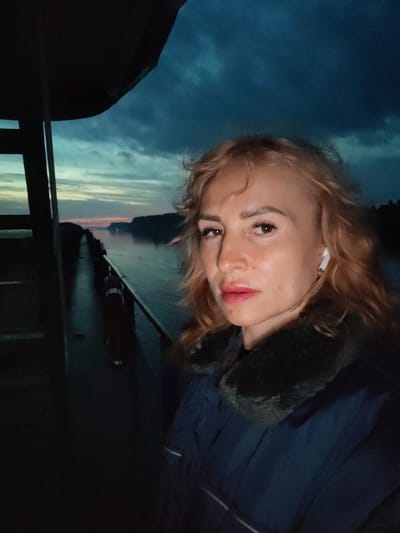 A woman with blonde hair and fair skin is taking a selfie from a balcony, likely on a boat, in the city of Badozhskoi Pogost, Russia. The sky is dark blue and cloudy, with streaks of light indicating a sunset or sunrise. A calm body of water stretches out in front of her, bordered by dark, tree-covered hills. The woman is wearing a dark blue coat with a fur collar and white earbuds. The overall mood of the image is serene and somewhat melancholic, due to the dim lighting and the vast, empty landscape.