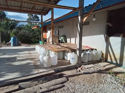 The image presents an outdoor scene in Ko Siboya, Thailand, during daylight hours. No individuals are present.
The foreground consists of a concrete slab with scattered wooden planks and construction debris, leading to a dirt and gravel area on the right. An elevated concrete platform in the mid-ground is covered with cardboard sheets, upon which numerous white, opaque plastic carboys, consistent with 5-gallon water bottles, are meticulously arranged in rows. At least one carboy displays a label with Thai text for "drinking water" (น้ำดื่ม) and a brand name, possibly "BOND."
To the left, a large blue plastic drum rests on the concrete, positioned in front of dense tropical vegetation featuring palm trees and other leafy plants, all under a blue sky with scattered white clouds.
On the right side of the image, a light-colored, single-story building with multiple horizontal glass-block-style windows and a blue roof gutter is visible. A screened window is situated on the far right. A white LED bulb hangs suspended from an overhead wooden beam of a partial roof structure that extends over the concrete platform and bottles. Against the building wall, a green tarp covers an unidentified object, and two green buckets are visible. A blue flexible pipe runs along the ground parallel to the building wall on the right. Strong shadows indicate bright sunlight originating from the left. The scene suggests a functional outdoor storage or processing area.