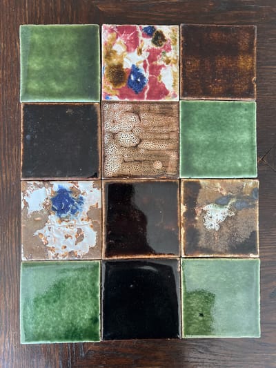 This media file displays a top-down view of twelve square tiles arranged in a 3x4 grid on a dark wooden surface. The tiles are varied in color and pattern, suggesting they might be decorative or part of a collection.

The top row features a dark brown textured tile on the left, followed by a solid green tile, a mottled brown and white tile with some blueish-gray inclusions, and another solid green tile on the right.

The middle row starts with a colorful tile featuring red, white, blue, and brown splotches. To its right is a tile with a distinctive speckled brown pattern against a lighter background. The next two tiles in this row are solid black, one appearing slightly glossy and the other more matte.

The bottom row consists of a solid green tile on the left, a solid black tile next to it, a tile with a white and brown base color overlaid with blue and brown abstract patterns, and finally another solid green tile on the far right.

The wooden surface beneath the tiles shows a visible grain pattern and appears to be a table or countertop. The lighting suggests an indoor setting, likely during daytime, as there are subtle reflections on some of the glossy tiles. There is no visible text in the image, and no people or specific activities are depicted. The context of Hämeenlinna, Finland, implies a potential cultural or artistic display.