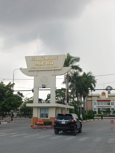 The image shows the Moc Bai International Border Gate in Ben Cau, Vietnam. A black SUV is stopped at a pedestrian crossing in front of the gate. The gate itself is a large, white archway with Vietnamese and English text reading "CUA KHAU QUOC TE MOC BAI" and "INTERNATIONAL BORDER GATE." Behind the gate, a building with the text "CONG HOA XA HOI CHU NGHIA VIET NAM" is visible. Palm trees line the road, and the sky is overcast. There are several people in the background, including one walking towards the building and others near the side of the road. A red and white stop sign is visible to the right of the SUV.