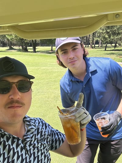 Two men in golf attire sit in a golf cart, holding drinks. The setting appears to be a golf course in Atizapán de Zaragoza, Mexico, evident from the green grass and trees in the background. The man on the top wears a blue patterned shirt and a gray cap. He holds a clear plastic cup filled with a dark liquid, possibly iced tea, in his gloved hand. The other man, facing the camera, wears sunglasses, a baseball cap, and a black and white patterned shirt. He also holds a cup with a straw. A segment of the yellow golf cart frame is visible.