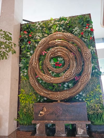 A verdant wall hosts a coiled, golden art piece composed of beaded bands forming concentric circles. The innermost circle cradles red and green foliage. The decorative sculpture rests upon a dark, rectangular plinth marked with circular and butterfly-shaped embellishments. Small plinths support the main structure, grounding the installation on a polished, reflective floor. Green plants also flank the base, softening the overall presentation.
