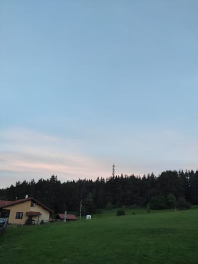 A panoramic, eye-level outdoor shot captures a rural landscape at dusk in Galabovo, Bulgaria. The sky dominates the upper portion, transitioning from a pale blue near the top to lighter shades with faint pink and orange hues closer to the horizon, indicating the setting sun. A dense, dark forest lines the middle ground, forming a backdrop for the scene. In the foreground, a lush green grassy field slopes gently downwards. On the left side of the field, a yellow house with a reddish-brown roof is visible, featuring windows and a small porch. To the right of the house, there is a smaller red-roofed structure, possibly a shed or outbuilding. A tall, slender radio tower stands prominently behind the forest line. Scattered throughout the field and at the edge of the forest are smaller trees and bushes. The lighting suggests a calm, quiet evening.