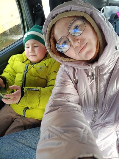 The image captures two individuals seated inside a vehicle, located in Старый Шагирт, Russia. In the foreground, an adult female with a light complexion is taking a selfie. She is wearing a light pink, quilted puffer jacket with the hood pulled up, revealing reddish hair or a scarf underneath. Black-framed eyeglasses rest on her face, reflecting a clear blue sky with white clouds. Her expression is neutral as she looks directly at the camera. To her left and slightly behind, a young child, also with a light complexion, is seated. The child wears a green and white striped knit beanie and a bright lime green puffer jacket with a visible circular logo on the left arm. Brown pants are also visible. The child is looking downward and to their left, holding a small, rectangular object in their left hand. The vehicle interior is dark-toned, with a black seatbelt mechanism visible near the window. Through the window, a blurred yellow surface and a partially discernible Russian license plate, "P700CA," are visible.
