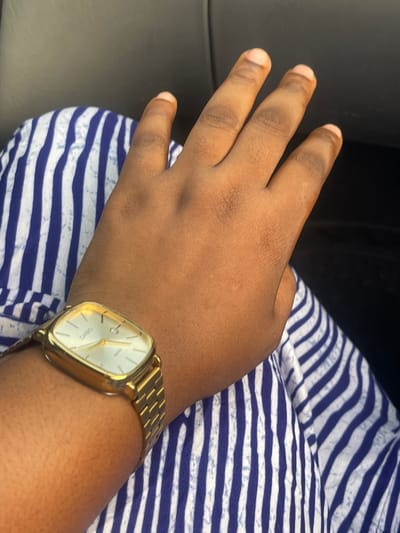 A person's left hand and wrist are shown resting on a surface with blue and white horizontal stripes. The hand is dark-skinned with visible fingers. A gold-colored wristwatch is on the wrist, displaying the time. The setting is indoors, near a dark-colored surface, possibly a car interior. It is impossible to ascertain if this image was taken in Kunkwaba, Nigeria or any other specific location.