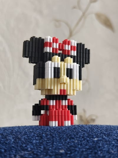 A close-up, eye-level, medium shot captures a three-dimensional, pixelated figure resembling Minnie Mouse constructed from small, interlocking plastic blocks. The figure is predominantly black, white, and red, with yellow accents for the face. It features large, black ears outlined with red and white stripes, a white face with black eyes and a red mouth, and a red dress with white accents. The figure stands on a textured, dark blue surface, possibly a rug or fabric. The background consists of a lightly textured, off-white wall with subtle shadows, suggesting it is indoors. The lighting is soft and diffused. There is no indication of the location being in Семилетка, Russia.