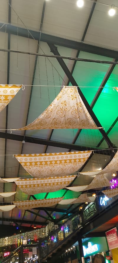 An upward shot captures a commercial space adorned with triangular fabric banners suspended from a metal ceiling structure. The banners feature a white background with golden floral and geometric patterns. Above the banners, the ceiling reveals metal beams and translucent green panels. Below, illuminated signs and strings of lights create a lively atmosphere, hinting at a vibrant marketplace or dining area.