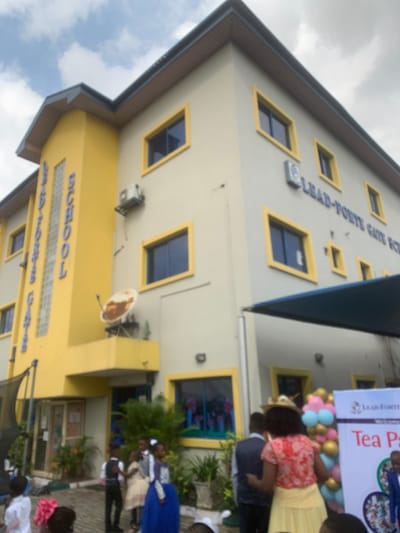 The image shows the exterior of Lead-Forte Gate School in Close, Nigeria. The school is located in a residential area with a paved driveway and a small garden in front of the building. It is a two-story building with a yellow and white facade, with several windows and a satellite dish on the roof. There is a sign on the wall that reads "Lead-Forte Gate School". The school is having a "Tea Party" event as a sign outside reads, "Tea Party". There are many children and adults present, some of them are dressed in their school uniforms, while others are wearing casual attire. There is a large inflatable archway and several balloons. The children are interacting with each other and the adults. There is a covered awning next to the entrance of the school. The weather is sunny, with a light overcast sky.