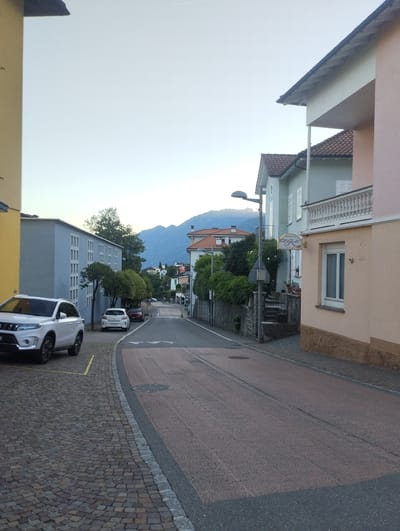 This image captures a street scene in Orselina, Switzerland, likely during the late afternoon or early evening given the soft, diffused light. The sky is a pale, clear blue, hinting at fair weather. The scene is dominated by residential buildings and a sloping road paved with asphalt and cobblestones.
On the left, a yellow building partially frames the view, with a white SUV parked on a cobblestone area marked by yellow lines. Adjacent to it is a light blue, multi-story building with many uniformly spaced windows. Further down the street, a white car is parked, and a darker car is visible behind it.
The road curves gently uphill, leading towards a backdrop of lush green trees and distant mountains. The architecture of the houses is varied, with some featuring terracotta tile roofs. One building on the right displays a sign that reads "Ristorante Al Gusto," indicating a restaurant. The building itself is painted in light pink and cream tones, with a balcony featuring white railings. Another building nearby is painted a pale blue-green.
The overall atmosphere is tranquil and residential, with no people visible in the frame, suggesting a quiet moment in the day. The presence of cars and buildings indicates an established urban or semi-urban environment. The mountains in the distance provide a scenic natural element to the otherwise built environment.
