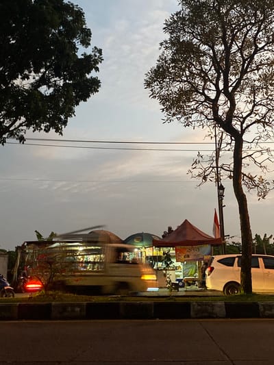 The image captures an outdoor street scene in the city of Parungserab, Indonesia, at dusk or early night. The foreground features a dark asphalt road, separated from the roadside by a low, dark-colored concrete median barrier.

In the mid-ground, a light-colored vehicle, likely a van or bus, is in motion from right to left, exhibiting significant motion blur. To the right of this moving vehicle, a white multi-purpose vehicle (MPV) is parked facing left. On the far left, a motorcycle with an illuminated red brake light is partially visible, also showing slight motion blur. Behind the median, a series of roadside commercial stalls are set up, illuminated by internal artificial light sources. These stalls include a dark, domed canopy structure and a distinct reddish-brown tent. Within the stalls, various items are displayed for sale. An Indonesian flag, red over white, is attached to a pole located behind the reddish-brown tent. A single human figure is faintly discernible standing near the reddish-brown tent.

The background consists of a sky with diffused, light-grey cloud cover. Two large trees frame the scene: one with dense, dark foliage on the left, and another with a more sparse, branched canopy on the right. Power lines are visible stretching horizontally across the upper portion of the frame. A dark, ornate street lamp pole stands partially obscured by the tree on the right.