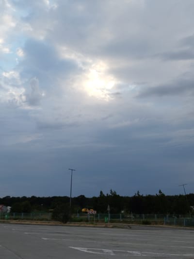 The image depicts an overcast sky with the sun partially obscured by clouds, casting a diffused light. In the foreground, there is a paved surface, likely a road or parking lot, with white lane markings visible. Beyond this surface, a line of dark green trees forms a dense border. A light pole stands tall on the left side of the scene. Further into the distance, various signs and what appear to be some yellow vehicles are present behind a fence. The overall impression is of an outdoor scene, possibly at the edge of a developed area, with natural elements contrasting with man-made structures. The location is identified as Tânganu, Romania.