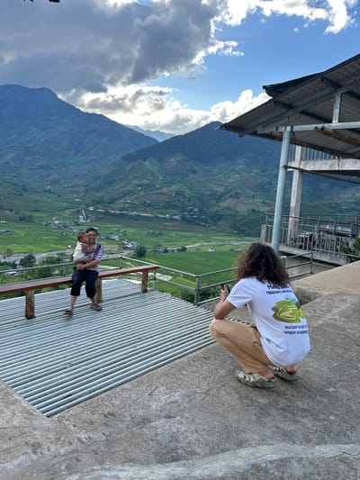 An outdoor scene captures a photographic activity at an elevated viewpoint overlooking a mountainous valley. The location is consistent with the Tú Lệ region of Vietnam, characterized by extensive terraced rice paddies.

In the mid-ground, an adult female is seated on a rustic wooden bench with metal supports, holding a young child. The woman wears a light-colored striped shirt, dark pants, and sandals. She is looking towards the camera's perspective with a slight smile. The child, wearing light-colored clothing, is held upright and also faces forward. They are positioned on a platform constructed of horizontal corrugated metal sheets.

In the foreground, on a concrete slab adjacent to the corrugated platform, an adult individual with long, curly brown hair is squatting. This person wears a white t-shirt with text and a graphic on the back, tan pants, and grey sandals, identified as Keen brand. The t-shirt text includes "MAO SERA TREKKING AND HOME..." and contact information ("WHATSAPP +44 34537177 Instagram: maosetrekking"). This individual is holding a black smartphone with both hands, aiming it at the woman and child on the bench, engaged in the act of photography.

The background features a vast landscape dominated by multiple layers of green, terraced rice fields descending into a valley. A river or stream meanders through the valley floor, and clusters of small buildings or villages are interspersed among the fields. Distant mountains frame the scene, their peaks partially obscured by a mix of dark and lighter clouds. The sky shows patches of blue to the right, with sunlight breaking through, while the upper left is heavily overcast. A metal railing is visible between the subjects and the expansive view. To the right of the frame, parts of an elevated structure with a metal roof, supported by white poles and beams, are visible, with bamboo elements integrated into its design. The concrete slab in the foreground exhibits minor surface cracks.