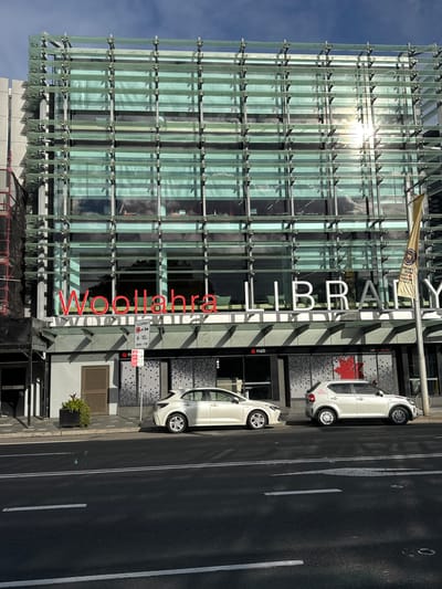 The photograph shows a modern glass-fronted building with the words "WOOLLAHRA LIBRARY" in large red and white lettering. The building appears to be in an urban setting, with a street and sidewalk in the foreground. Two white cars are parked on the street in front of the building. The sun is shining brightly, reflecting off the glass facade and creating a glare. A flagpole with a yellow banner is visible to the right of the building. The building's architecture is characterized by a grid-like structure of glass panels and metal framing. The sidewalk features a planter with greenery and a grey door with horizontal slats. A street sign with parking restrictions is also visible. The overall impression is of a well-maintained public building in a contemporary urban environment.